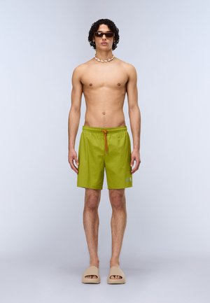 Mand uden skjorte med krøllet hår iført mørke solbriller, skalsmykke, limegrønne shorts med orange snøre og beige slidesandaler.