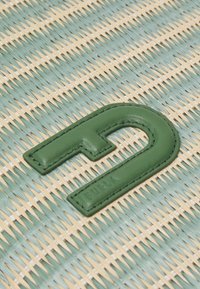 Zielona skórzana litera "D" na plecionej niebiesko-żółtej tkaninie, z teksturowanym wykończeniem i wyraźnymi detalami na szwach.