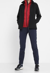 Personne portant une veste noire zippée sur une polaire rouge, un pantalon slim bleu marine et des chaussures de sport noires avec des semelles blanches, debout les mains dans les poches.