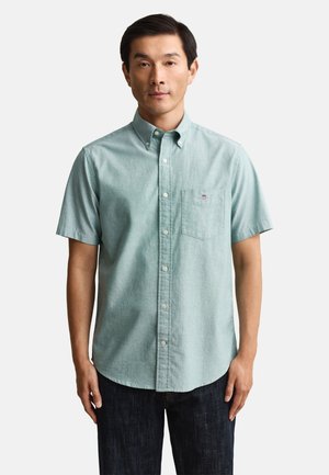 Man die een lichtgroen overhemd met korte mouwen en borstzak draagt, staand tegen een effen witte achtergrond.