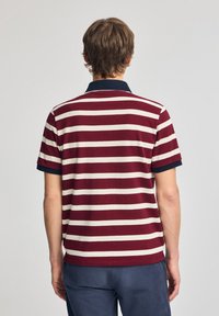 Pikétröja i vinröd med horisontella krämfärgade ränder. Har en marinblå krage och korta ärmar. Tillverkad av bomullstyg.