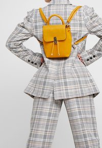 Żółty skórzany plecak o uporządkowanym kształcie, z zapięciem na klamrę i regulowanymi paskami, noszony na kraciastej marynarce z różnymi wzorami krat.