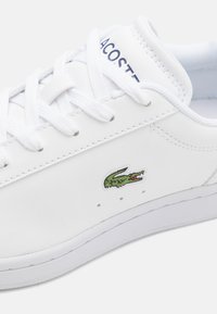 Fehér bőr sneaker, kerek orral, lapos fehér fűzőkkel, perforációkkal a légáteresztésért, és hímzett zöld krokodil logóval az oldalon.