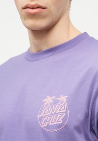 Νεαρό άτομο που φοράει μπλουζάκι skateboard Santa Cruz σε λιλά απόχρωση με ροζ λογότυπο φοίνικα στο στήθος, ορατό από τον λαιμό έως το άνω μέρος του κορμού.