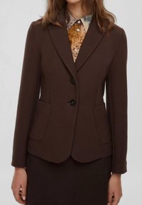 Blazer marrone texturizzato con due tasche frontali e due bottoni neri. Indossato sopra una camicia a motivi multicolori. Stile con vestibilità sartoriale.