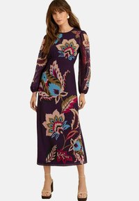 Maxime dress in tumman violetti, jossa on värikkäitä kukkakuoseja. Pitkät läpinäkyvät hihat ja istuva vyötärö. Yhdistetty vaaleanruskeisiin platform-sandaaleihin.