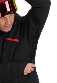 Giacca nera con accenti rossi, chiusura con zip, fodera in rete sotto le ascelle e logo sulla manica. Il materiale sembra resistente e flessibile.