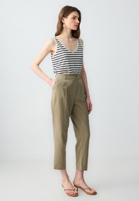 Pasiasty top na ramiączkach w kolorze kremowym i granatowym, zestawiony z wysokimi spodniami w jasnym khaki. Model stoi z rękami w kieszeniach, nosząc proste sandały.