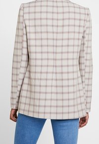 Veste beige à carreaux avec une coupe ajustée, présentant des carrés bleus et marron, des manches longues et une finition en tissu lisse.