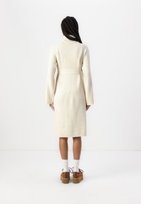 Rochie crem, tricotată cu nervuri, cu mâneci lungi, talie cu brâu și lungime midi, asortată cu pantofi cu platformă maro și sosete albe.