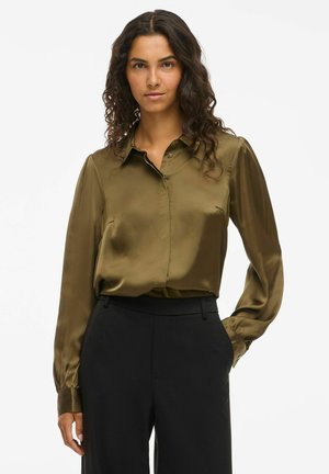 Blusa in satin verde oliva con colletto a bottoni, maniche lunghe e polsini. Texture liscia, design minimalista. Abbinata a pantaloni neri.