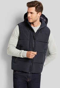 Marineblaue Steppweste mit Kapuze, vorn mit Reißverschluss und zwei seitlichen Taschen. Darüber wird ein hellgrauer Pullover mit gestepptem Muster getragen.