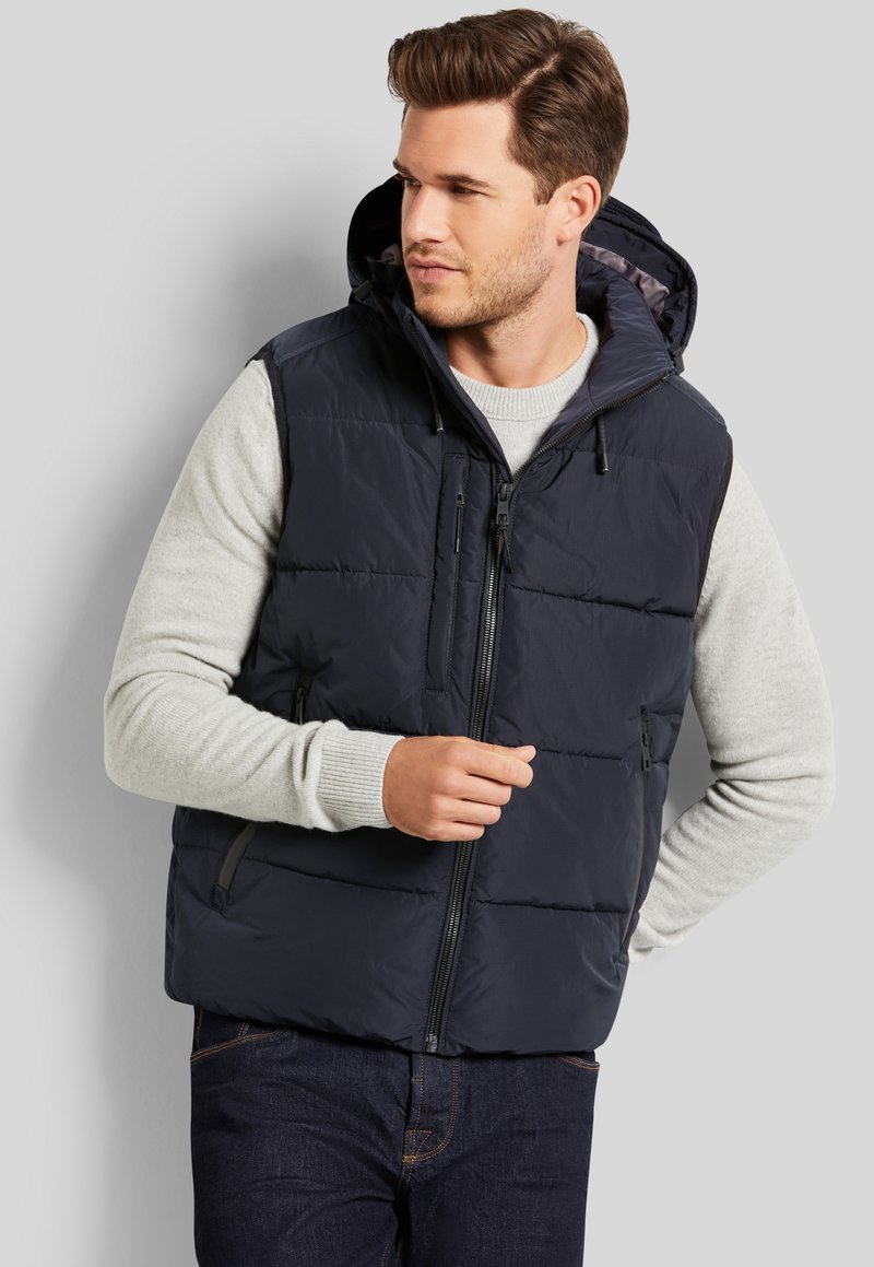 Marineblaue Steppweste mit Kapuze, vorn mit Reißverschluss und zwei seitlichen Taschen. Darüber wird ein hellgrauer Pullover mit gestepptem Muster getragen.