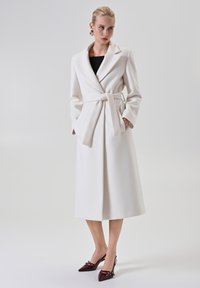 Donna che indossa un lungo cappotto bianco con cintura sopra un top nero, abbinato a tacchi a punta slingback di un borgogna scuro, in piedi di fronte a uno sfondo neutro.