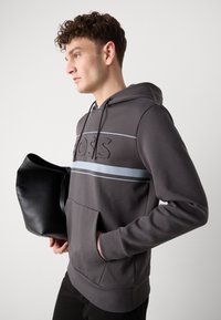 Felpa grigia con logo testurizzato, strisce orizzontali in grigio scuro e azzurro chiaro, e tasca frontale. Borsa nera tenuta nell'altra mano.