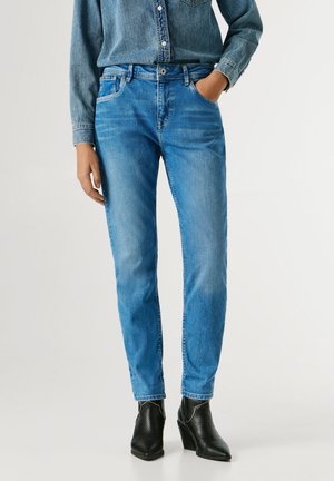 Personne portant un jean en denim bleu, une chemise en denim rentrée, avec une main dans la poche et des bottines noires texturées à talon.