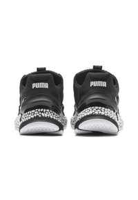 Baskets de sport noires avec une semelle texturée, présentant un talon noir brillant, un logo blanc et un motif distinctif de taches blanches sur la semelle.