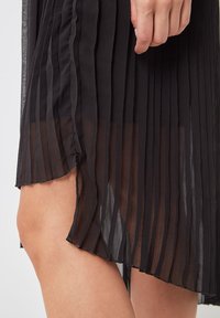 Falda negra plisada con tejido transparente, presenta rayas verticales y un dobladillo ligeramente curvado. Textura ligera y fluida.