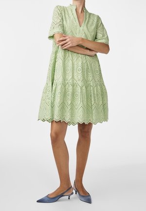 Osoba ubrana w jasnoróżową koronkową sukienkę z oczkami o długości do kolan z falistym dołem i niebieskie, szpiczaste, zakładane na piętę buty na niskim obcasie, ramiona skrzyżowane.