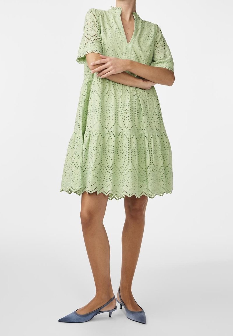 Personne portant une robe vert clair en dentelle à œillets longueur genou avec un ourlet festonné et des escarpins slingback bleus pointus à petits talons, bras croisés.