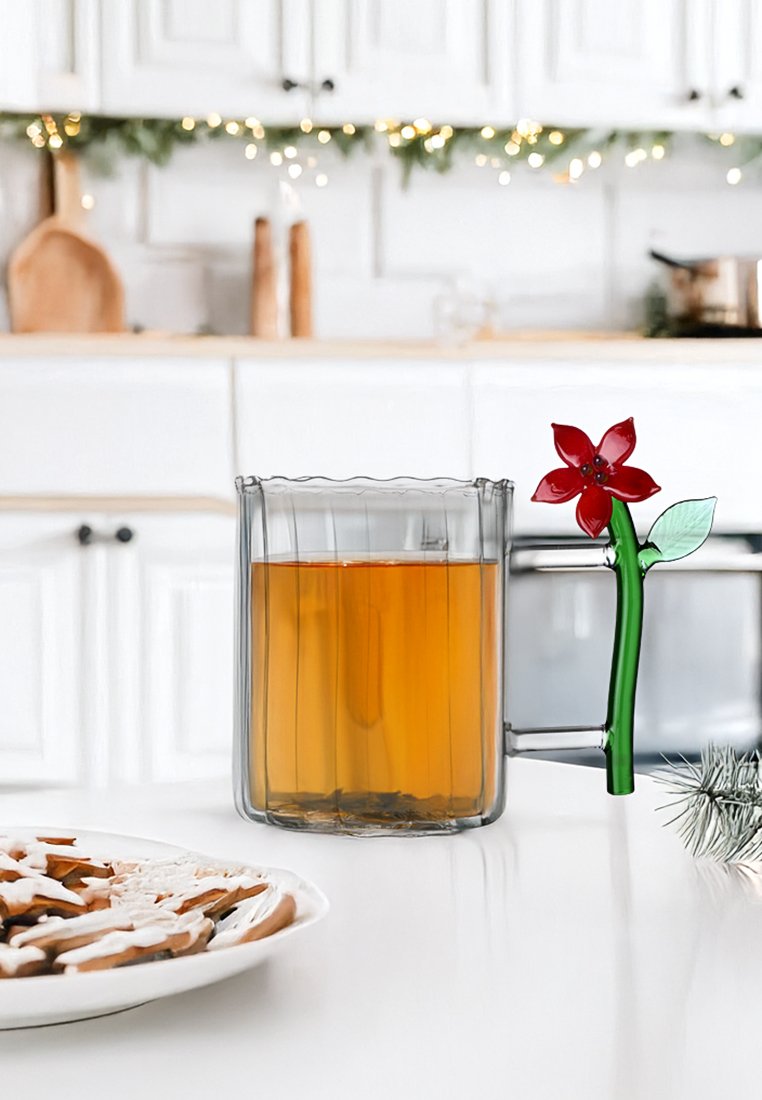 Przezroczysty szklany kubek o falistych brzegach, wypełniony bursztynowym płynem, z zielonym uchwytem w kształcie łodygi kwiatu, ozdobionym czerwonym kwiatem.