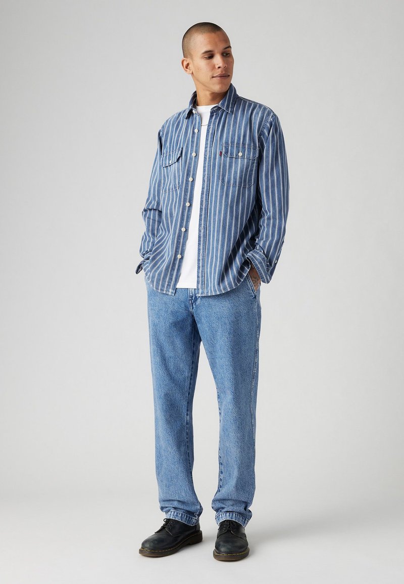 Blau-weiß gestreifte Button-Up-Bluse, Jeans und schwarze Schuhe. Auffällige Details sind zwei Brusttaschen und eine lockere Passform.