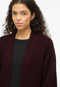Vīnsarkanā adītā kardigana ar atvērtu rāvēju, teksturētu rakstu un nolaistām plecu līnijām. Valkāta virs melnas krekla, ar brīvu piegriezumu.