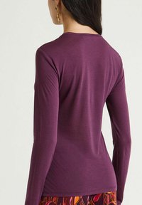 Langärmeliges Oberteil in Tiefviolett mit rundem Ausschnitt und glatter Textur, kombiniert mit gemustertem, buntem Stoff am Saum.