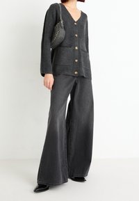 Šedý žebrovaný cardigan s V-výstřihem a dvěma předními kapsami, zapínání na knoflíky, spárovaný se širokými šedými džínami a černými špičatými botami.