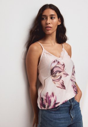 Donna con lunghi capelli scuri che indossa una canottiera floreale rosa chiaro e jeans blu, in piedi contro una parete bianca liscia, che guarda di lato.