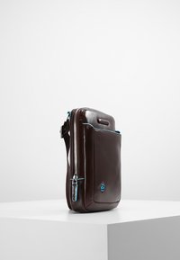 Borsa in pelle marrone con una texture liscia, dotata di chiusura a zip, accenti blu e una tasca frontale. Forma rettangolare compatta.