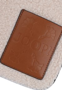 Tessuto beige texture con una toppa in pelle marrone stampata con "JOOP!" e motivi decorativi. Bordo nero attorno alla toppa.