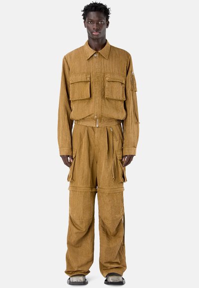 Persona alta con chaqueta cargo marrón holgada y pantalones anchos a juego con múltiples bolsillos, de pie mirando hacia adelante.