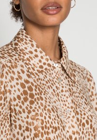Camicia beige con stampa leopardata marrone, dotata di colletto e patta a bottoni, realizzata in materiale liscio con una leggera texture lucida.