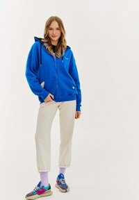 Królewsko-niebieska bluza z zamkiem na całej długości, z kieszeniami z przodu i detalem z logo. Połączona z kremowymi spodniami i wielokolorowymi trampkami.