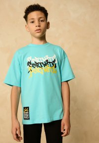 Tricou turcoaz cu mânecă scurtă, cu text grafic multicolor pe față și o etichetă neagră cu detalii aurii în formă de coroană la margine.