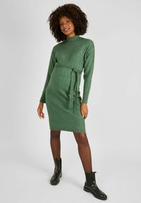 Abito di maglia verde con collo alto, vita cincta e maniche lunghe. Indossato con stivaletti neri alla caviglia. Tessuto testurizzato con un motivo mélange.