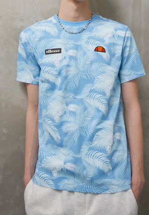 Osoba w jasnoniebieskiej koszulce z białymi wzorami palmowych liści, naszywkami logo ellesse, srebrnym naszyjniku i jasnoszarych spodniach.