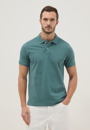 Mann, der ein türkisfarbenes Polohemd mit kurzen Ärmeln und weiße Hosen trägt, steht mit einer Hand in der Tasche vor einem einfarbigen hellen Hintergrund.