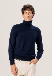 Marineblauer Rollkragenpullover aus weichem Strickmaterial, taillierter Schnitt mit langen Ärmeln und geripptem Saum. Kombiniert mit hellen Hosen.