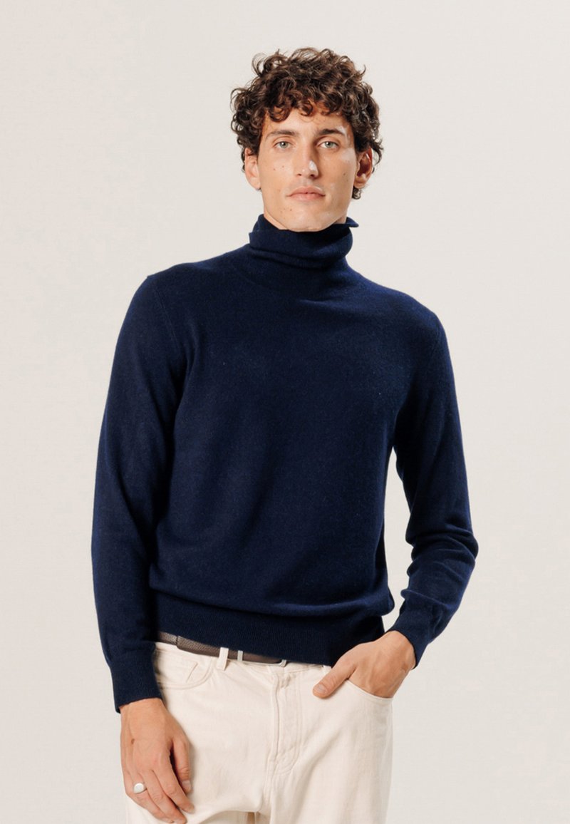 Marineblauer Rollkragenpullover aus weichem Strickmaterial, taillierter Schnitt mit langen Ärmeln und geripptem Saum. Kombiniert mit hellen Hosen.