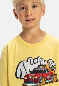Camiseta de algodón amarilla con un gráfico de un coche rojo con llamas, contornos en negro y texto en negrita. Mangas cortas y escote redondo.
