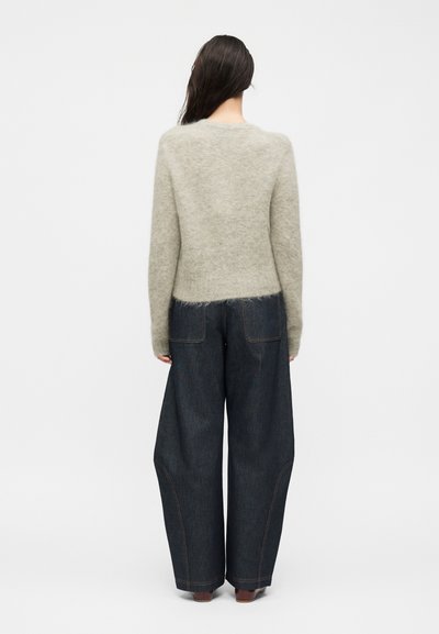 Femme aux longs cheveux foncés portant un pull gris clair duveteux et un jean large en denim foncé, debout dos à un fond blanc uni.