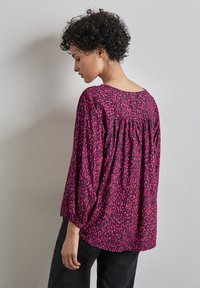 Luźna różowa bluzka z czarnym abstrakcyjnym wzorem, okrągłym dekoltem i marszczonym tyłem. Długie rękawy z elastycznymi mankietami.