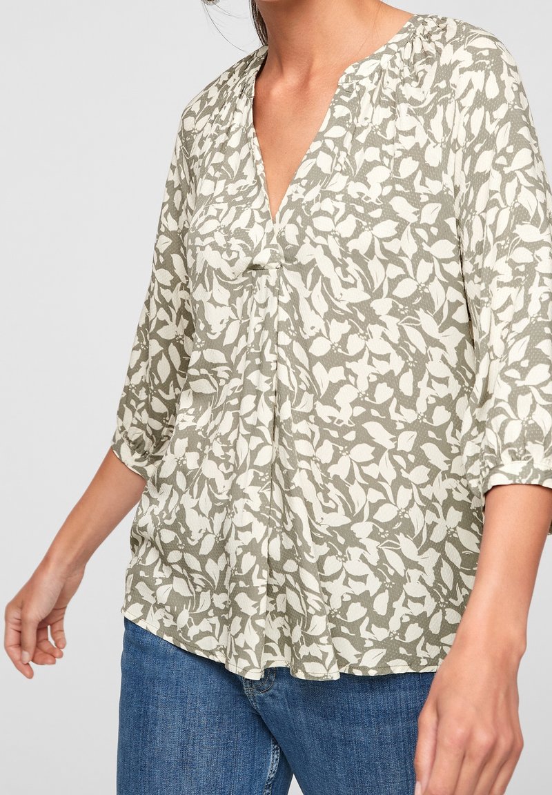 Femme portant un chemisier ample à manches longues avec un motif abstrait de feuilles blanc et vert olive, associé à un jean bleu.