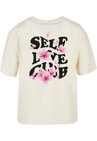 Krémové tričko s krátkym rukávom s nápisom "SELF LOVE CLUB" v tučnom čiernom písmene, ozdobené ružovými kvetinovými detailmi na chrbte.