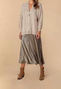 Beige Cardigan mit fransigen Kanten, silbernen Knöpfen und zwei Taschen, kombiniert mit einem glänzenden, olivgrünen Midirock und braunen Stiefeletten.