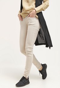 Vaqueros skinny beige con una textura suave, dos bolsillos frontales y un ajuste de cintura media, combinados con un chaleco largo negro y zapatillas deportivas negras.