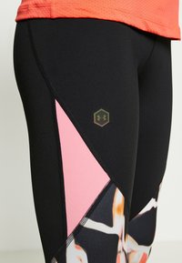 Černé sportovní legíny s růžovými akcenty, které mají geometrický vzor a malé logo v hexagonu na boku. Hladká, elastická tkanina.