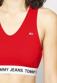 Raudona gumbinė trumpa palaidinė su giliu V formos iškirpimu, kuri turi baltą juostą su juodu raštu "TOMMY JEANS" ir mažą logotipą priekyje.
